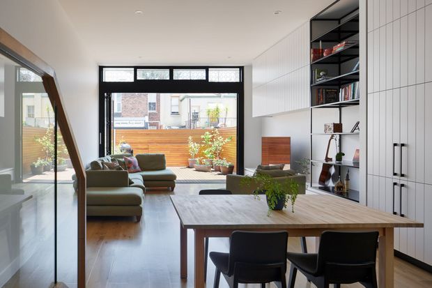 Bright open-plan contemporary interior combining a dining area and living room. A substantial timber dining table with dark upholstered chairs occupies the foreground, while a sofa and armchair sit beyond near full-height black-framed glazing opening toward a timber-fenced courtyard. White vertical wall panelling, integrated cabinetry, open shelving, timber flooring, and restrained decor create a clean, refined urban home setting.