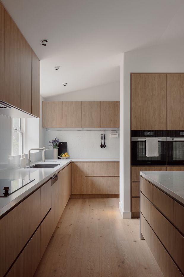 A contemporary galley-style kitchen with warm light-oak cabinetry extending along both sides, integrated black ovens, pale stone countertops, a stainless sink and tap, and a central island. The room has white walls and ceiling, light timber flooring, recessed downlights, and a clean, highly tailored built-in layout. Natural light enters through a side window.