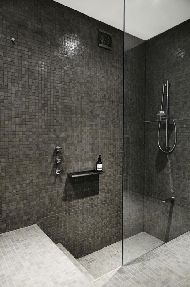 A contemporary walk-in shower enclosed by a clear frameless glass screen. The walls are clad in small dark grey mosaic tiles, while the shower floor uses lighter grey mosaic tiles. Chrome controls, a handheld shower, recessed shelf, and ceiling ventilation grille are visible. The space has a dark, moody, minimalist appearance.