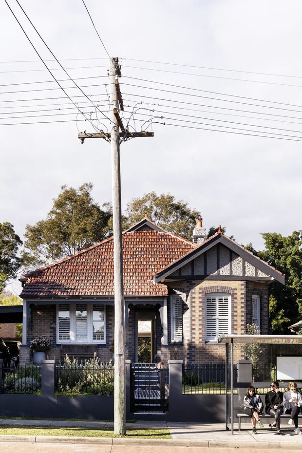 Street-facing view of a traditional brick bungalow with a terracotta tiled hipped roof, front gable, decorative brickwork, white plantation-style shutters, chimney, front garden, and low rendered boundary wall. Utility poles and overhead wires dominate the foreground, with a bus shelter and people visible at the street edge.
