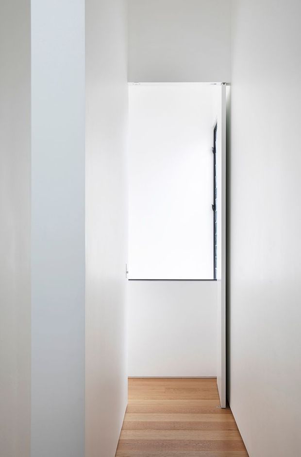 A narrow, minimalist white hallway or passage leading to a bright window or glazed opening. The space has smooth white walls and a pale timber floor, with a simple white door or panel at the end and a slim dark window frame visible along its edge.