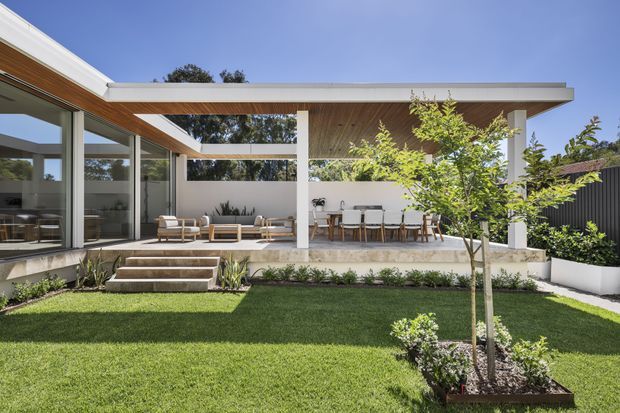 Bright contemporary single-storey home viewed from a landscaped backyard, featuring a broad flat roof with white structural columns, extensive floor-to-ceiling glazing, warm timber-lined soffits, a covered outdoor dining and lounge area, and steps leading to a manicured lawn and garden.