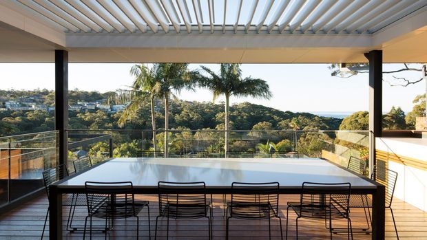 Covered outdoor dining terrace with a long white dining table and black wire-frame chairs, enclosed by glass balustrades and framed by dark structural posts. A slatted pergola roof provides shade, with expansive views over leafy suburban hills and distant water.