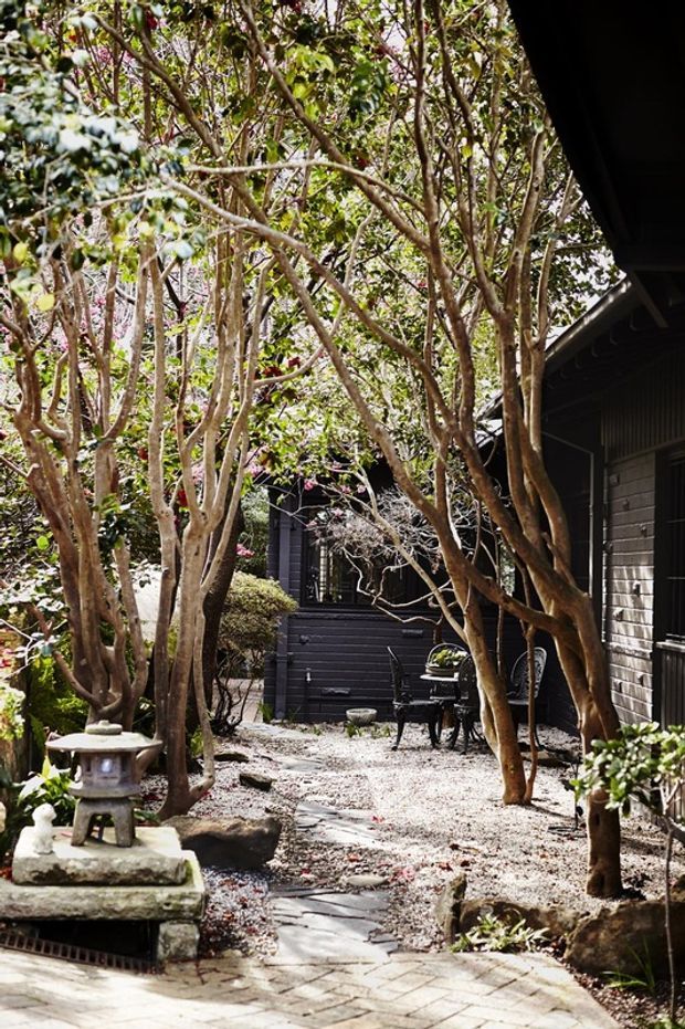 A tranquil, enclosed courtyard garden beside dark-painted timber-clad buildings. The space features mature multi-trunk trees, gravel groundcover, stepping stones, sculptural rocks, clipped shrubs, and a small Japanese-style stone lantern. A compact outdoor seating area is partially visible in the background, creating a shaded, intimate garden setting.