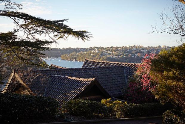 Exterior view across a landscaped garden toward a traditional dark tiled pitched roof, with a leafy waterfront suburb and bay visible in the distance. The image is dominated by the roofline, surrounding mature vegetation, and an elevated coastal setting.
