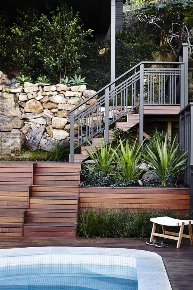A landscaped outdoor pool area on a sloped site, featuring terraced timber decking and planter beds, a natural stone retaining wall, timber stairs with grey metal balustrades, dense planting, and a tiled swimming pool in the foreground.