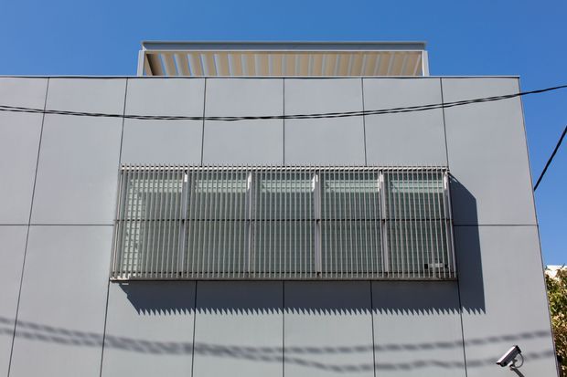 Exterior architectural photograph of a modern, rectilinear building facade clad in large pale panels. A projecting upper structure has a slatted soffit, while the central window opening is screened by dense vertical metal fins that cast strong shadows. Overhead utility wires cross the facade, and a security camera is visible at the lower right.