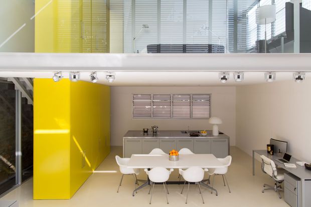 A bright, double-height contemporary open-plan interior combining a dining area, compact kitchen, and home office. A glossy yellow feature wall and matching upper-level wall add a bold accent, while white furniture, pale flooring, extensive horizontal blinds, and exposed track lighting create a clean modern workspace-like setting. A mezzanine or upper-level walkway overlooks the dining area.