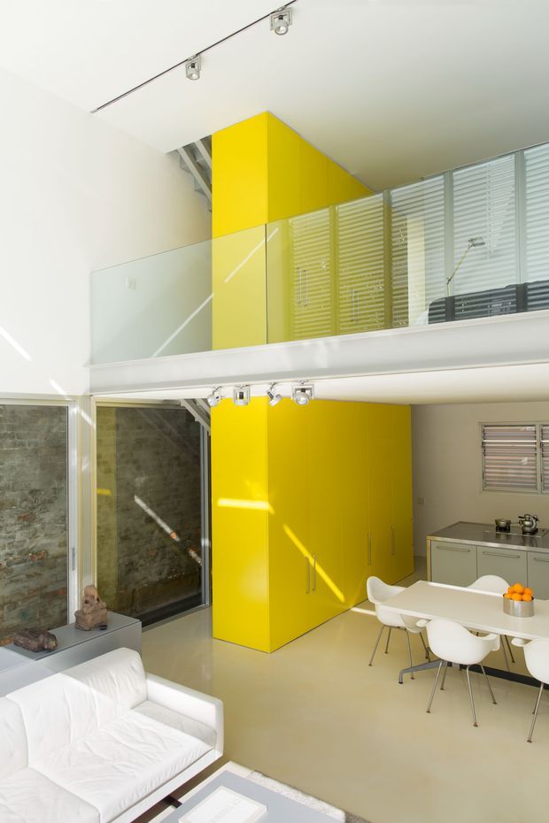 Bright double-height open-plan interior with a mezzanine level and glass balustrade. The main living area contains a white sofa and dining table with white chairs, while a vivid yellow built-in cabinetry volume rises through both levels. A compact kitchen with pale cabinetry sits beside the dining area. Exposed stonework is visible at the rear, with horizontal blinds, track lighting, and strong sunlight creating a crisp contemporary atmosphere.
