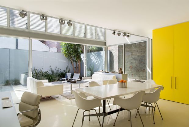 Bright open-plan contemporary interior combining a dining area and living room. A glossy white dining table with sculptural white chairs sits beside a white sofa and lounge seating. Full-height glazing, high-level louvred windows, exposed pale brickwork, polished light flooring, track lighting, and a vivid yellow built-in cabinet create a clean, colourful modern space with a courtyard garden visible beyond.