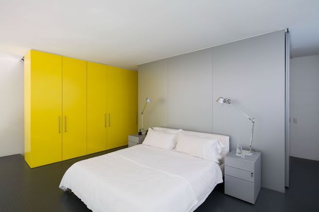 A minimalist bedroom with a low-profile white bed, white bedding, slim bedside tables, articulated reading lamps, and a large bright yellow built-in wardrobe along the left wall. The room has pale grey wall panels, a smooth white ceiling, and a dark floor, creating a clean high-contrast modern interior.