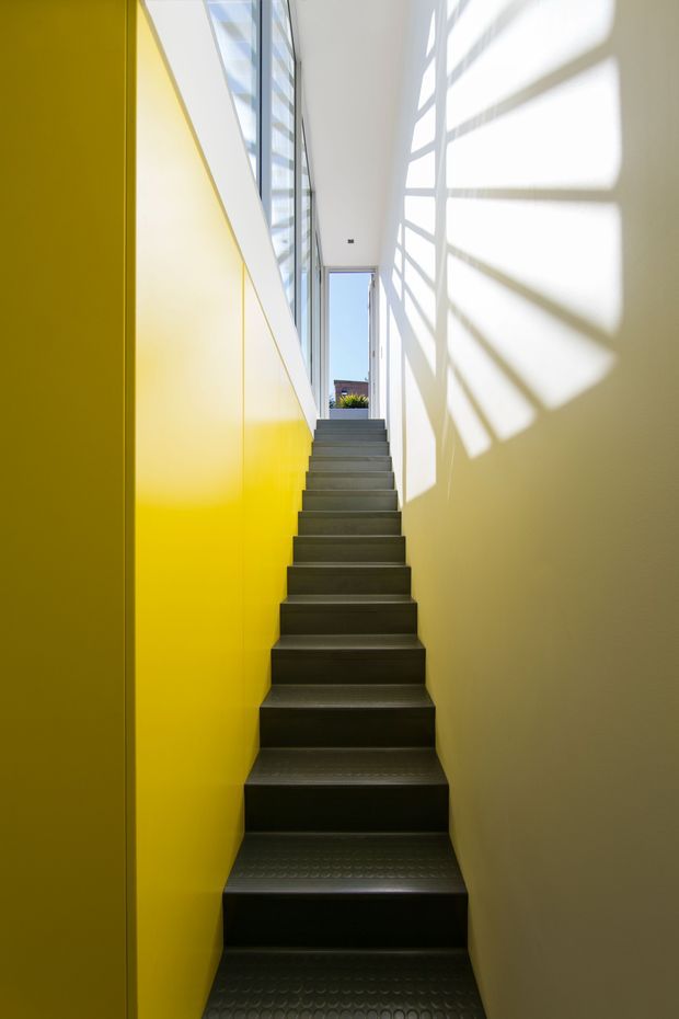 A narrow, steep interior stairway rises toward a bright landing and tall glazed opening. The stair treads are dark with a subtle patterned finish, flanked by a vivid yellow built-in wall on the left and a plain light wall on the right. Strong sunlight through the upper glazing casts dramatic geometric shadows across the right wall, creating a bold, graphic atmosphere.