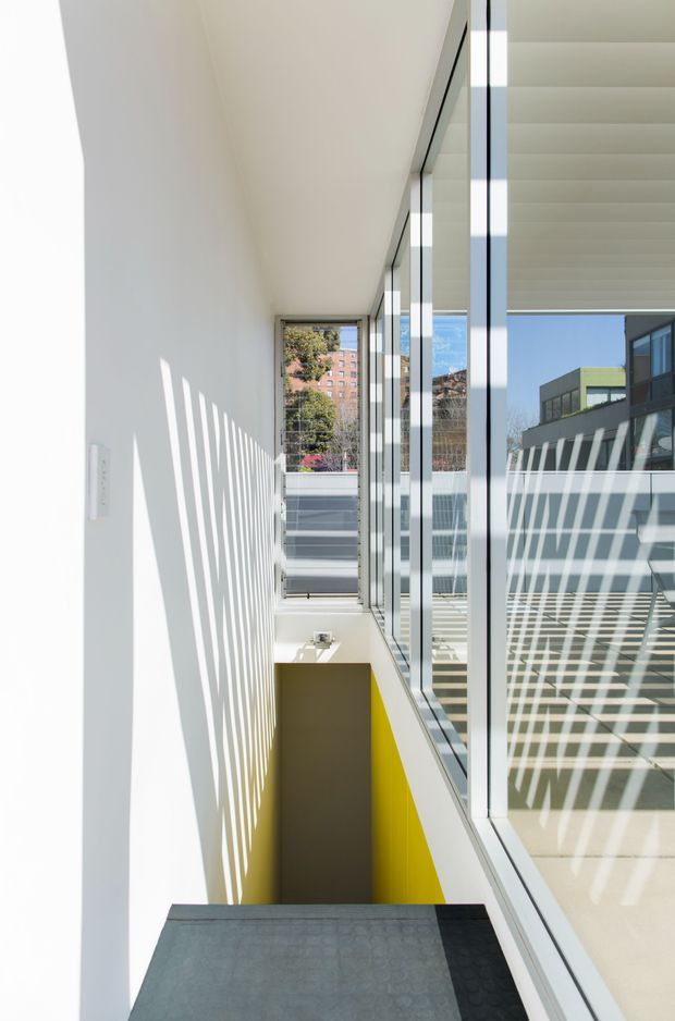 A bright, narrow interior circulation space beside full-height glazing, with strong linear sunlight and shadow patterns across white walls. A stair descends to a lower level with a vivid yellow-painted side wall, while reflections reveal adjacent modern building forms outside. The space has a crisp, minimalist architectural character and abundant daylight.