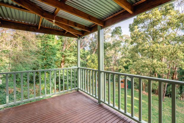 Covered outdoor deck or verandah overlooking a leafy wooded garden. The space has timber decking, pale green painted railings and posts, and a corrugated metal roof supported by exposed timber beams.