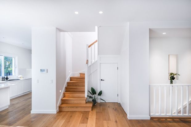 Bright contemporary residential interior showing a white-painted entry hall with warm timber flooring, a central timber staircase with white balustrades, and a closed door beneath the stairs. A white kitchen is visible to the left and an open stair landing or void with matching railing is visible to the right. Recessed ceiling lights and minimal furnishings create a clean, airy appearance.