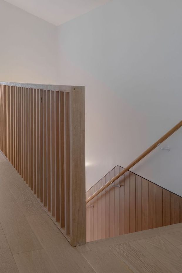 Interior view of a minimalist stairway and landing with pale timber flooring, vertical timber balustrade slats, timber wall panelling, and a simple rounded timber handrail against white walls. The stairwell has a clean, warm, understated contemporary appearance.