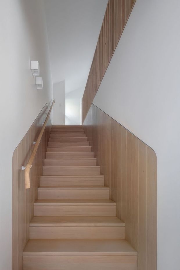 A minimalist interior stairway with light timber stairs, matching vertical timber wall panelling and a simple timber handrail. White walls and ceiling create a bright, clean, sculptural composition, with the stair enclosure narrowing toward a softly lit upper landing.