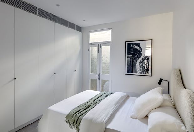 A bright, compact contemporary bedroom with a neatly made bed, white bedding, and a muted sage-green throw. Full-height white built-in wardrobes line the left wall, incorporating a linear ventilation grille near the ceiling. A glazed door and upper window bring in natural light, while a black wall-mounted reading lamp and framed architectural photograph provide restrained accents.