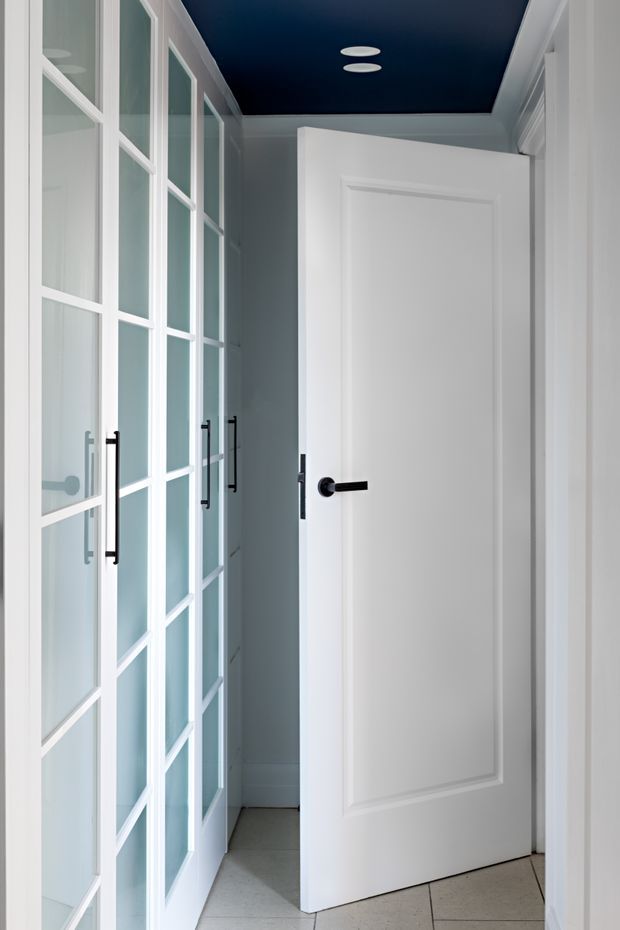 Bright, narrow interior hallway or entry passage with white traditional panelled doors, black lever hardware, full-height white-framed glazed cabinetry or partition doors along the left side, pale tiled flooring, and a contrasting deep navy-blue ceiling with recessed circular lights. The space has a crisp, refined, high-contrast transitional feel.