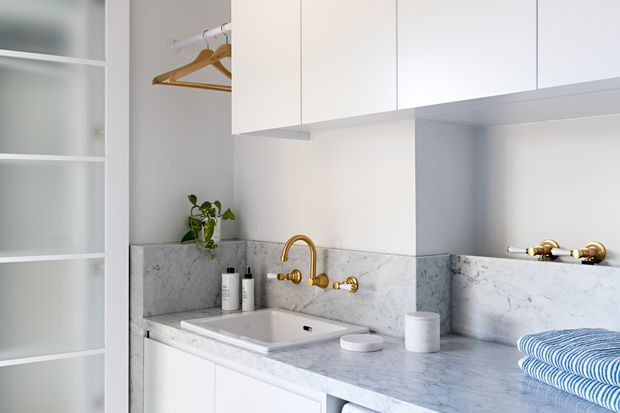 Bright, compact laundry room with white overhead and lower cabinetry, open shelving, a hanging rail, and a pale grey-white marble or stone benchtop with matching splashback. A deep inset sink with polished brass wall-mounted tapware is set into the counter. The space has a clean, refined look with a small trailing plant and folded blue-striped textiles.