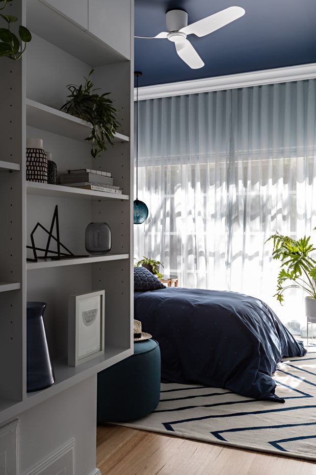 A stylish bedroom viewed past a built-in open shelving unit. The room features a dark navy bed and bedding, sheer floor-to-ceiling curtains, indoor plants, a geometric area rug, light timber flooring, and a deep blue ceiling with a white ceiling fan. The shelving displays minimal decorative objects and books, creating a refined contemporary interior with strong navy-and-white contrast.