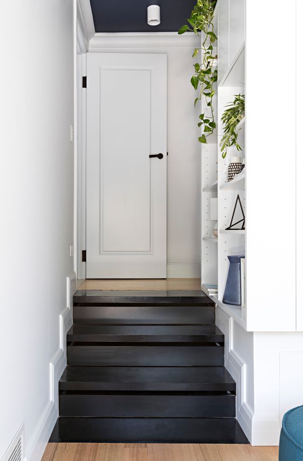A narrow, bright residential stairway with dark painted timber steps leading to a white paneled interior door. White walls, decorative wall mouldings, a dark charcoal ceiling with a simple cylindrical pendant light, and built-in white shelving with plants and decorative objects create a refined, compact transitional interior.