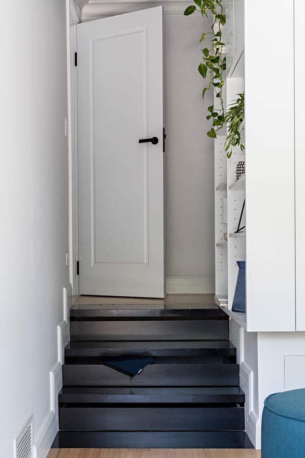 A narrow interior stairway with dark painted timber treads and risers leading up to a white paneled door. The walls, skirting boards, door, and decorative crown moulding are white, creating a bright contrast with the staircase. Built-in white shelving and cabinetry line the right side, displaying books and trailing plants, with part of a blue upholstered seat visible in the foreground. A small floor vent is set into the left wall.