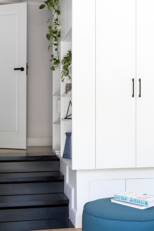A bright, refined residential hallway and stair landing with a white paneled door, built-in white storage cabinetry, open display shelving with plants and decor, and dramatic dark-painted stairs. A blue upholstered ottoman occupies the foreground. The space combines crisp white joinery and trim with black stair treads and a blue accent piece.
