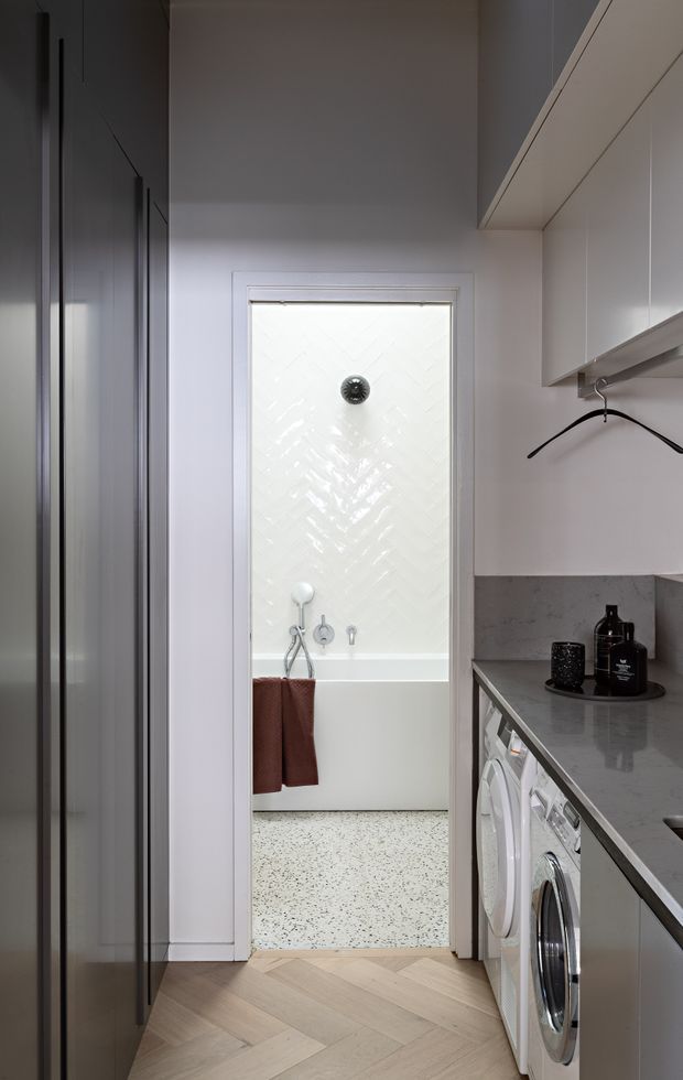 A compact, contemporary laundry viewed toward an adjoining bathroom. The laundry has tall dark built-in cabinetry on the left, white overhead cabinetry on the right, a dark grey stone benchtop and splashback, and front-loading washing machines beneath the counter. Light timber herringbone flooring leads into the bathroom, which features a freestanding or built-in white bath, white textured wall tiles, and a terrazzo-style floor.