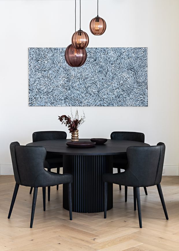 A refined dining room with a round black dining table featuring a fluted cylindrical pedestal base, surrounded by four dark upholstered dining chairs. Three smoked amber glass pendant lights hang above the table, while a large blue-grey abstract textured artwork provides a feature wall backdrop. The room has white walls with subtle lower wall moulding and a pale timber parquet floor.