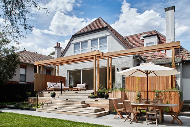 Rear exterior view of a renovated multi-level house with a steep terracotta tiled roof, pale rendered walls, dormer windows and a tall chimney. A timber pergola extends across a broad elevated timber deck with steps, built-in planting, outdoor seating and a dining area beneath a large umbrella. The setting includes a small lawn and landscaped backyard, with a bright, partly cloudy sky.