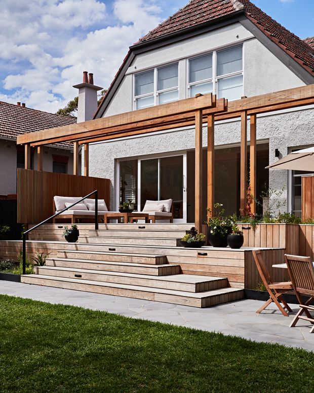 A renovated residential rear facade with a broad timber deck and multi-level stepped outdoor terrace. The light textured exterior walls, pitched terracotta-tile roof, white-framed windows and glazed doors sit behind a timber pergola structure. The terrace includes built-in timber seating or planters, outdoor lounge furniture, potted plants, a pale stone-paved area and lawn.
