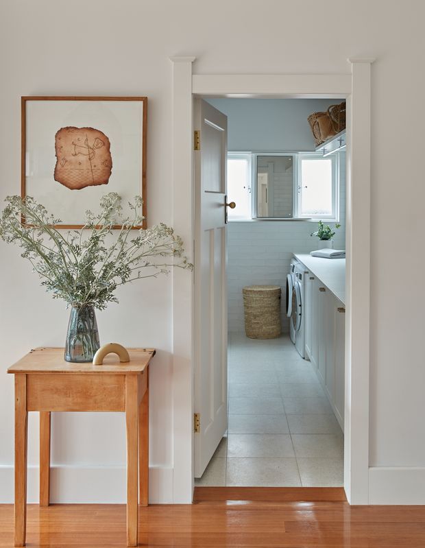 A bright interior view from a refined hallway or entry area into a compact laundry room. The foreground has warm timber flooring, white painted walls with classical trim, a timber console table, framed artwork, and a vase of foliage. Through the open paneled door, the laundry features white cabinetry, a pale tiled floor, white wall tiling, front-loading appliances, a woven basket, and a small window. The space has a calm, clean, light-filled character.