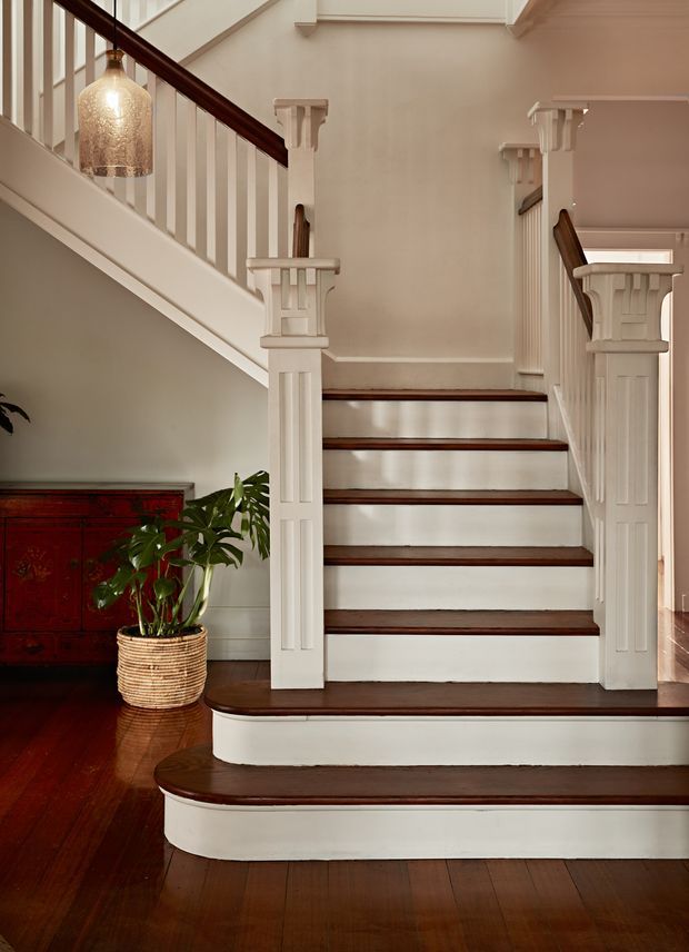 A refined residential stairway with dark stained timber treads and handrails, white painted risers, balustrades and decorative newel posts. The staircase has a curved lower step and rises to a landing, with warm beige walls, polished dark timber flooring, a textured pendant light, and a red-toned timber sideboard with a potted plant. The detailing gives the space a heritage-inspired, traditional character.