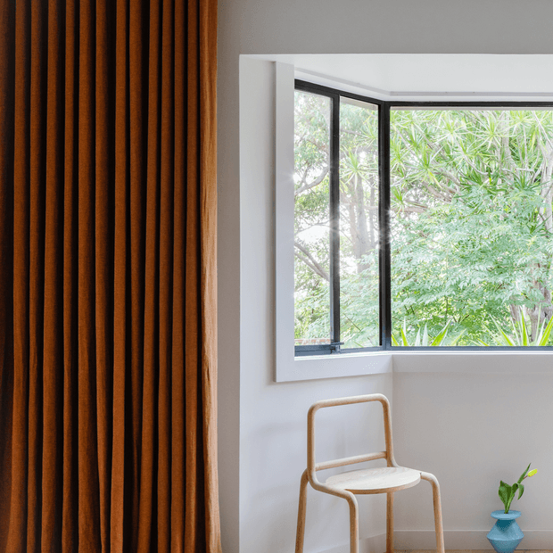 A bright, minimalist interior corner featuring a large black-framed corner window overlooking lush greenery. A warm rust-brown vertically pleated curtain occupies the left side, while a simple light timber chair and small blue geometric planter sit against the white wall.