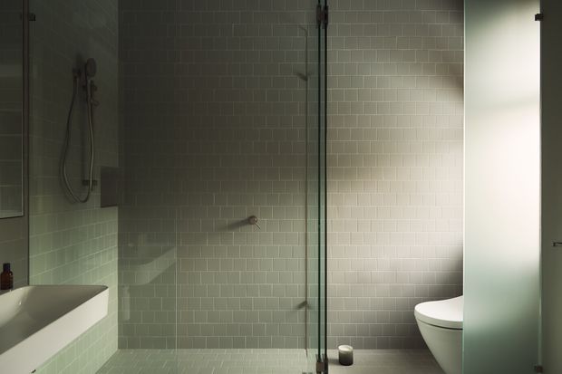 A contemporary bathroom with a walk-in shower enclosed by clear glass, pale green-grey subway wall tiles, matching small-format floor tiles, a white freestanding or integrated bathtub at left, and a white toilet partly screened by a frosted glass panel at right. The space is dimly lit with a focused warm light on the tiled shower wall and dark metal shower and glass hardware.