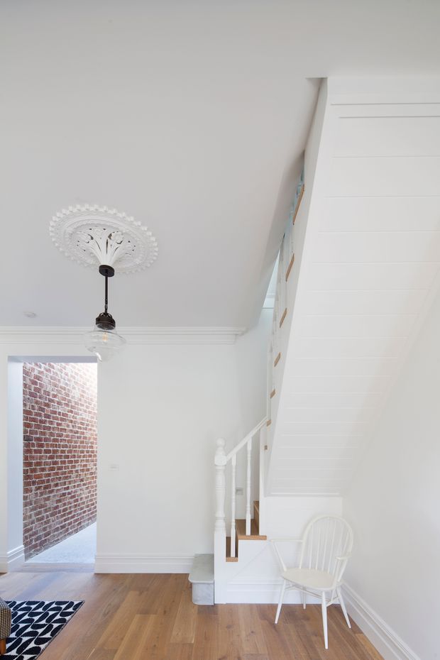 Bright white residential hallway and stairway with traditional detailing, including ornate ceiling medallion and pendant light, decorative cornices, white-painted timber stair balustrade, and a white paneled sloping ceiling beneath the stairs. Warm timber flooring, a patterned rug, and a white chair add contrast. An exposed red-brick wall is visible through the open doorway.