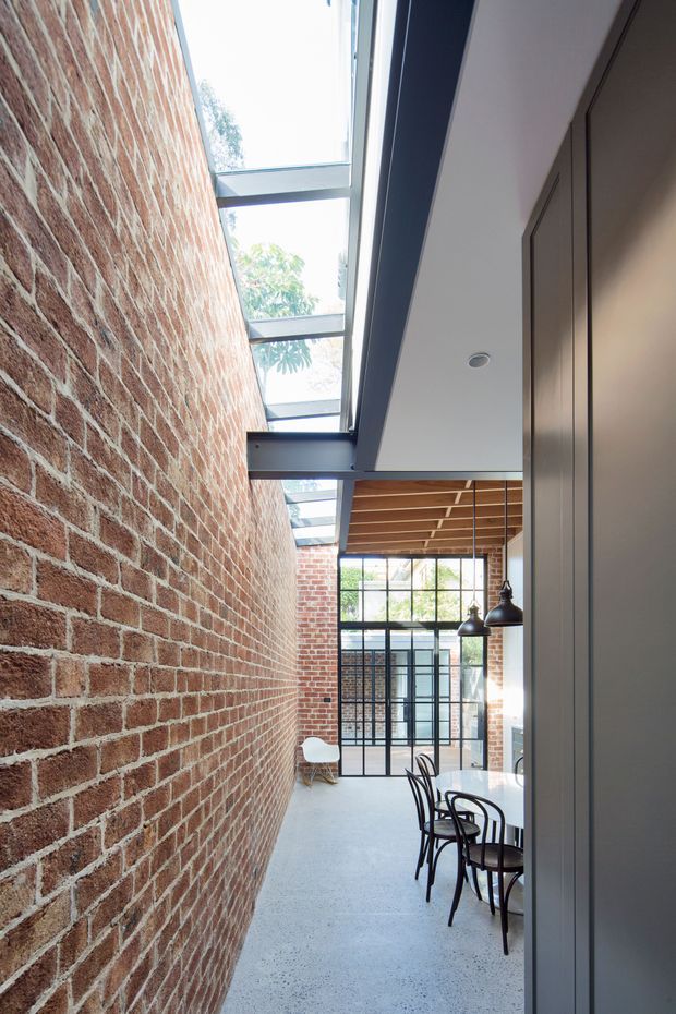 A narrow, light-filled contemporary interior with an exposed red brick wall, polished concrete floor, expansive steel-framed glazing, and a linear skylight. A dining area with dark chairs and pendant lights sits beneath a warm timber slatted ceiling, with tall dark built-in cabinetry along the right wall.