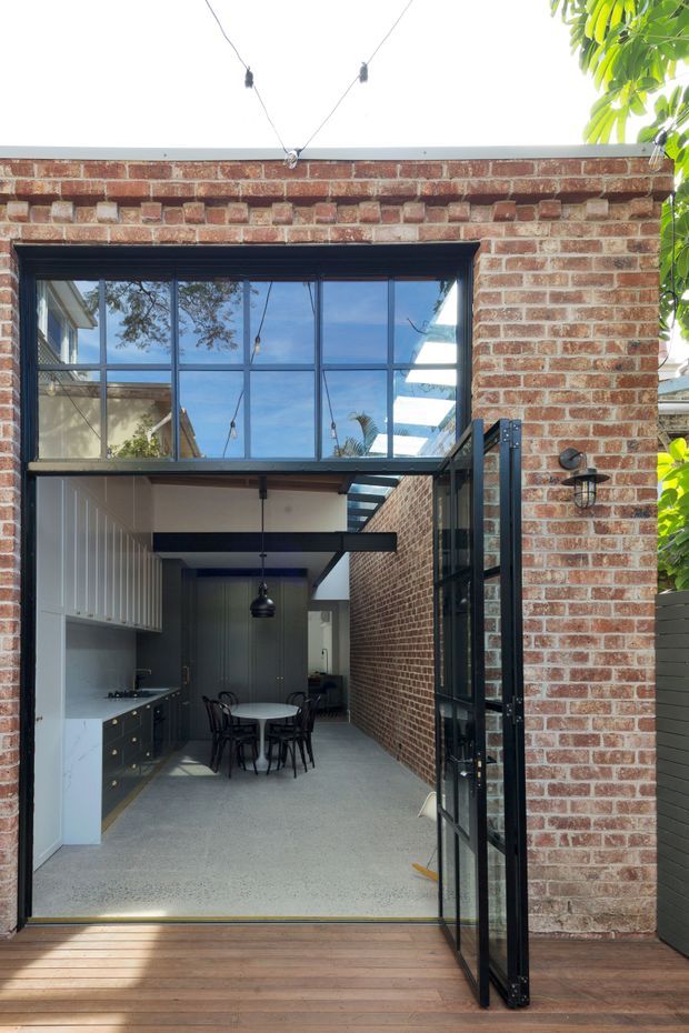 View from a timber deck into a narrow, courtyard-like extension with exposed red brick walls, black-framed glazed doors and a large clerestory-style window. The interior contains a compact kitchen and dining area, with dark cabinetry, a light stone or terrazzo-like floor, and a small round dining table. A glazed overhead connection brings abundant natural light into the space.