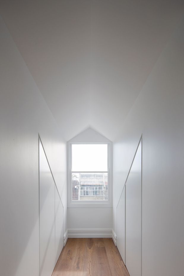 A narrow, minimalist hallway or landing with white built-in cabinetry on both sides, a pitched ceiling forming a pointed alcove around the rear window, white trim, and warm timber flooring. The window provides daylight and a view of a neighbouring building.