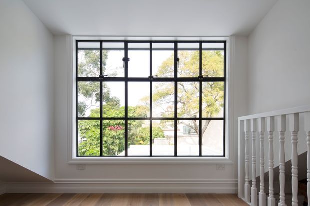 Bright, mostly empty interior landing or stair hall with a large black-framed multi-pane window overlooking leafy greenery. White walls, ceiling, and traditional skirting frame warm timber flooring, while a white turned-baluster stair railing occupies the right side.