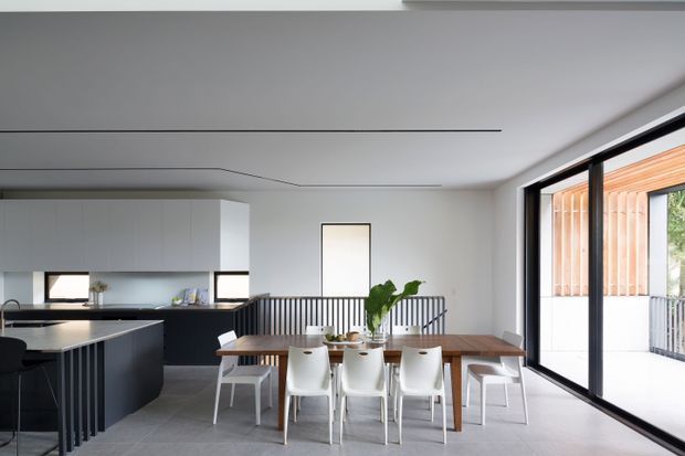 A bright, contemporary open-plan kitchen and dining area. The space features a large dark kitchen island with integrated seating, white flat-panel cabinetry, a long timber dining table with white chairs, light grey tiled flooring, and expansive black-framed sliding glass doors opening to a covered outdoor area. A black metal stair or balustrade element and concealed linear ceiling lighting add architectural definition.