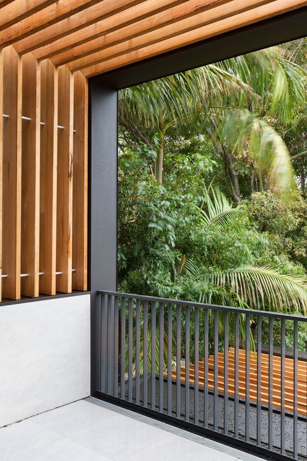 A covered outdoor balcony or terrace framed by warm timber rafters and vertical timber screening, with dark metal balustrades, pale tiled flooring, and views into dense tropical garden vegetation. The space has a contemporary indoor-outdoor character with strong timber detailing and a sheltered, elevated feel.