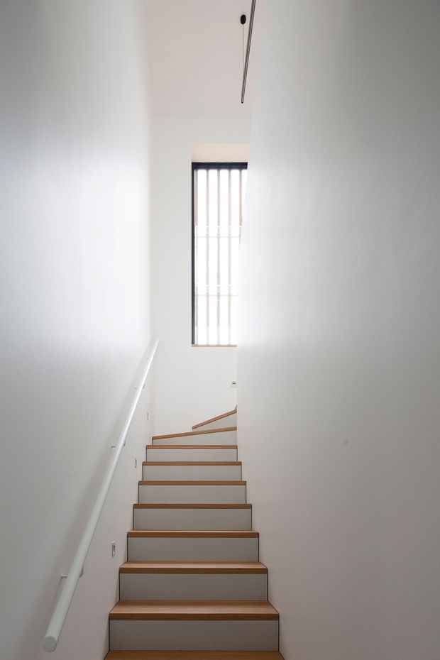 A narrow, minimalist stairway viewed from below, with white painted walls, a white handrail, and a straight flight of stairs with warm timber treads and pale risers. A tall vertical window with dark framing and slim vertical bars brings daylight into the stairwell.