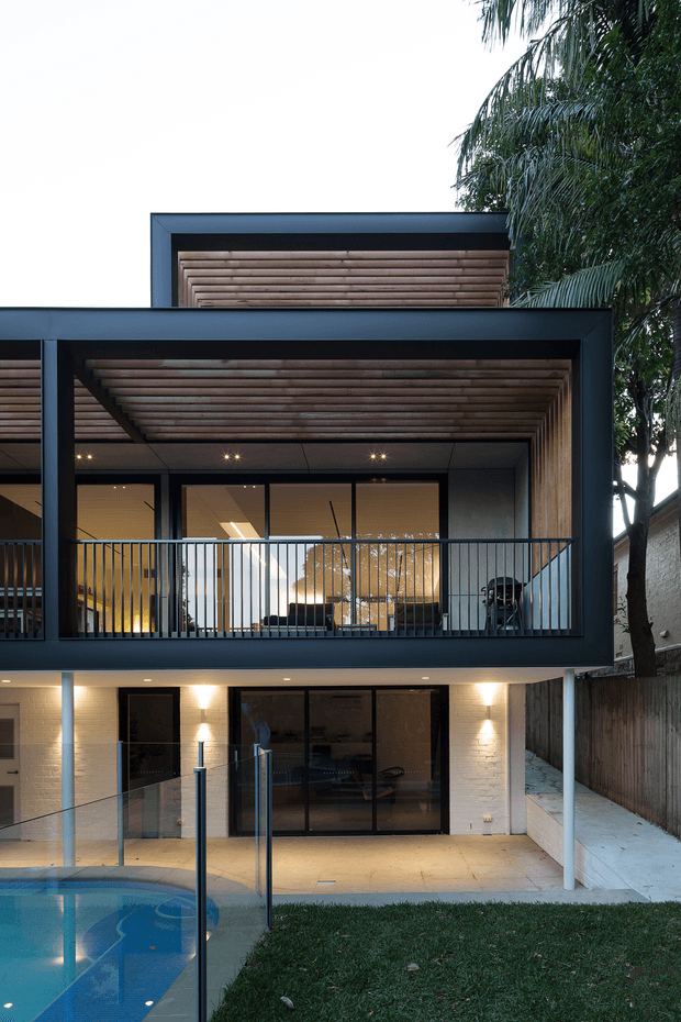 A contemporary multi-storey residential facade viewed from the backyard, featuring a deep upper-level covered balcony with a timber-lined ceiling, dark metal framing, glass doors, and slim black balustrades. The lower level has pale brick walls, large glazed doors, and warm exterior lighting, opening onto a small lawn and swimming pool. Mature palms and boundary fencing frame the property.