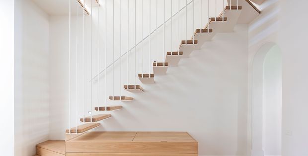 Bright minimalist interior featuring a sculptural floating timber staircase with slim white vertical balustrades, rising above a built-in timber-clad platform or storage volume. White plaster walls, a tall arched opening, and abundant natural light create an airy, refined composition.