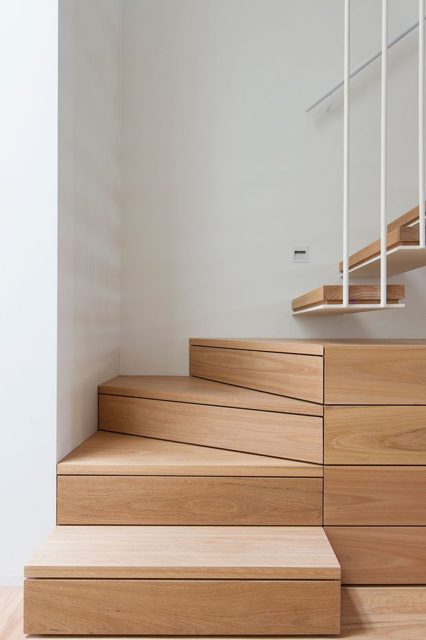 Minimalist interior view of a compact staircase with integrated timber storage drawers and stepped cabinetry. The stairs and joinery are finished in light natural wood, set against smooth white walls, with slim white balustrade rails and a clean, space-efficient design.