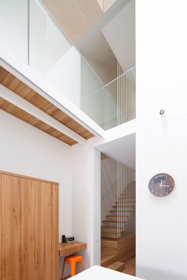 Bright, vertically oriented interior view of a modern multi-level home entry and stair hall. White walls and structural planes surround warm timber flooring, a tall timber door or built-in joinery, timber ceiling slats, and an open staircase with floating timber treads and slim white balustrades. A glazed upper-level balustrade and wall-mounted clock are visible, with a small timber console and orange stool in the lower entry area.