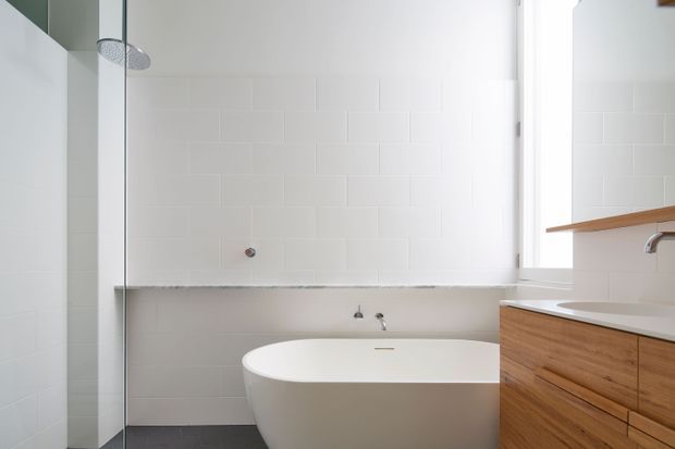 Bright contemporary bathroom featuring a freestanding white bathtub beneath a tiled wall, a frameless glass shower screen, and a timber vanity with a white countertop. A large window and open wall shelving add natural light and storage.