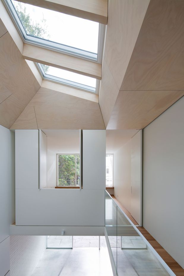 A bright, minimalist interior circulation space viewed upward through a tall void. Pale timber plywood cladding forms the ceiling and upper walls around two elongated skylights, while white walls, concealed joinery, glass balustrades and polished concrete surfaces create a clean, modern architectural composition. Windows and a mirrored or glazed opening reveal adjacent areas and greenery outside.